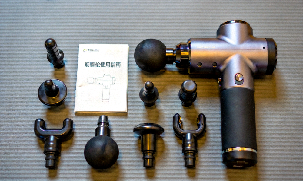 多种型号筋膜枪与配套按摩头在瑜伽垫上排列展示，旁边放着使用说明书